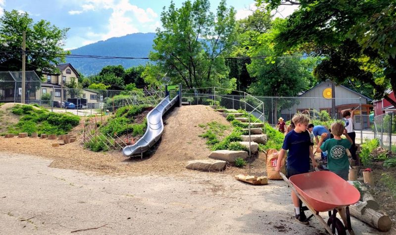 Part of the new playground. A slide goes down a dirt bank. Plants growing around. Children pushing a wheelbarrow on the side.