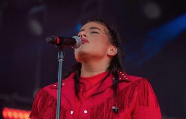Shawnee Kish performing on stage. She is wearing a red jacket and singing into a microphone. There is a dark blue and black background.