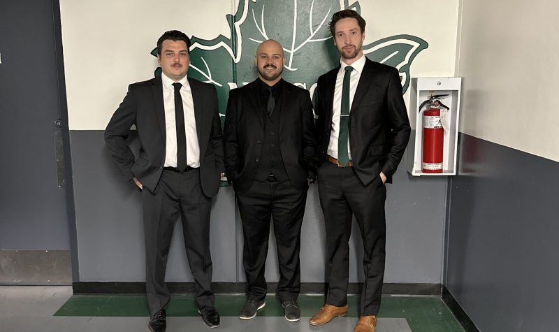 Three men in suits stand in front of a wall with the Nelson Leafs logo on it.
