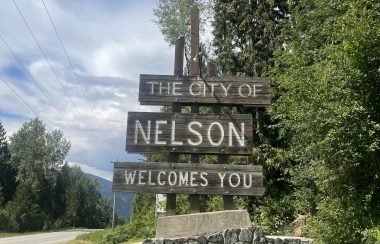 A wooden three board sign mounted on top of a fieldstone base and sitting beside a paved road reads 'The City of Nelson Welcomes You'. Trees and blue skies in the background.