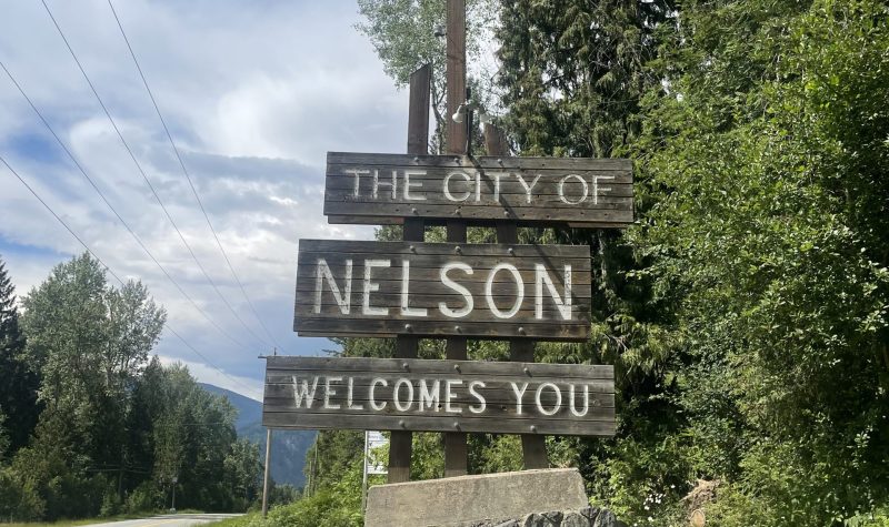 A wooden three board sign mounted on top of a fieldstone base and sitting beside a paved road reads 'The City of Nelson Welcomes You'. Trees and blue skies in the background.