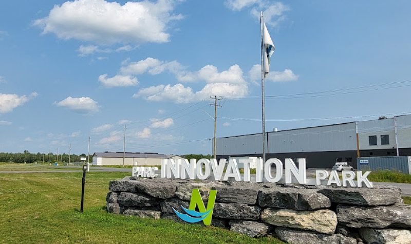 A white sign sits on squared limestone blocks with a white industrial building behind and green grass around it. The sign says Parc Innovation Park.