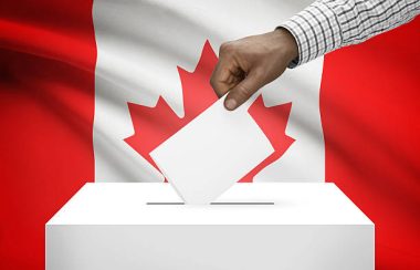 Le bras d'un homme noir, portant une chemise carreautée, dépose un carnet de vote dans une boite blanche, devant le drapeau canadien.