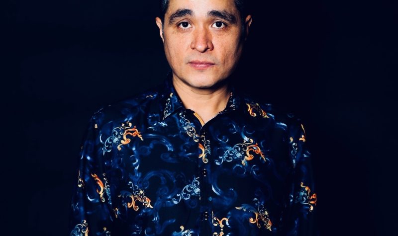 A man wearing a dark blue shirt with a multicoloured pattern poses in front of a dark blue backdrop.