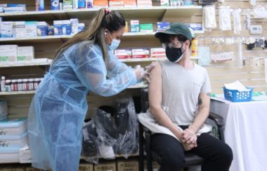 A person is sitting down while a person standing gives them a needle shot inside a clinic