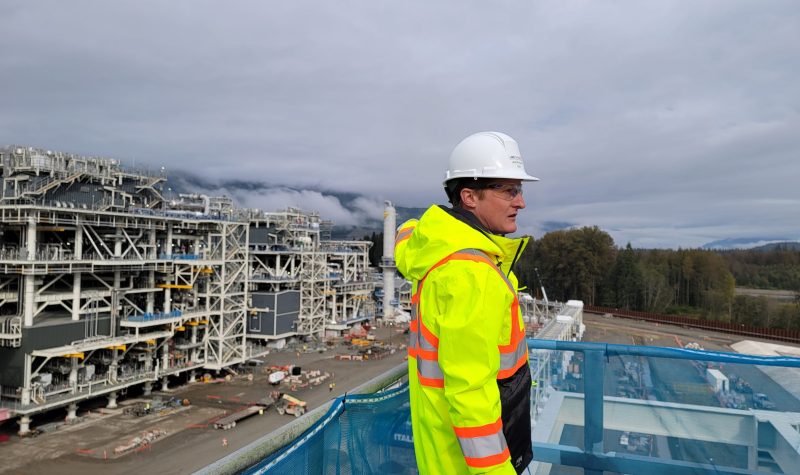 A man in a yellow high visibility jacket and white hard hat standing in front of industrial park