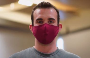 A still of John Ferguson wearing a red facemask and grey shirt.