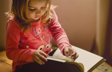 On voit une petite fille d'environ 4 ans qui est assise par terre et qui feuillète un livre. La petite fille a les cheveux blond et porte un chandail rose.