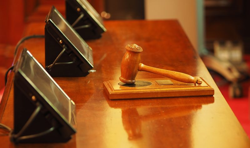A brown judge's gavel sits on its block on a brown table in a courtroom. Three black monitors are sitting infront of the gavel and block.