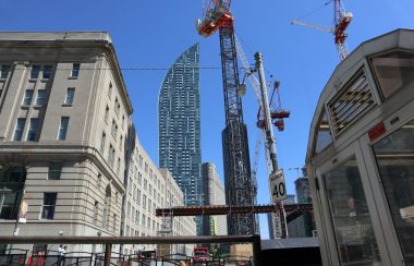 Large cranes with buildings around. The sky is sunny and blue.