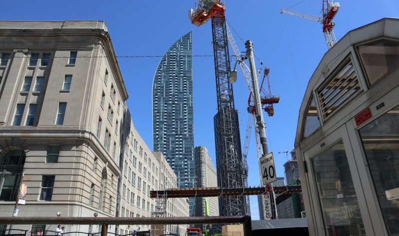 Large cranes with buildings around. The sky is sunny and blue.