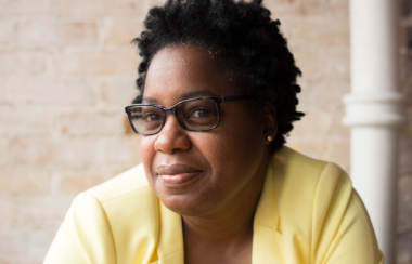 Wearing a yellow blazer and black rimmed glasses, with a brick wall in the background, Dr. Kathy Hogarth is sitting for a headshot picture.