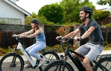 On peut voit un homme et une femme âgés d'environ 30 ans faire du vélo électrique sur une route d'un quartier résidentiel. Les deux cyclistes portent un casque et semblent avoir du plaisir.