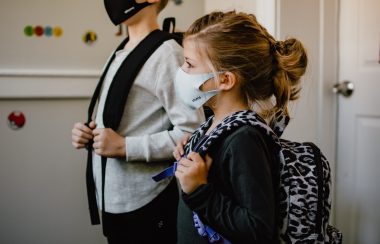 Two children, one taller than the other, both wear health masks and their backpacks in a classroom.