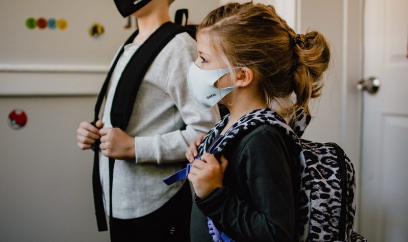 Two children, one taller than the other, both wear health masks and their backpacks in a classroom.