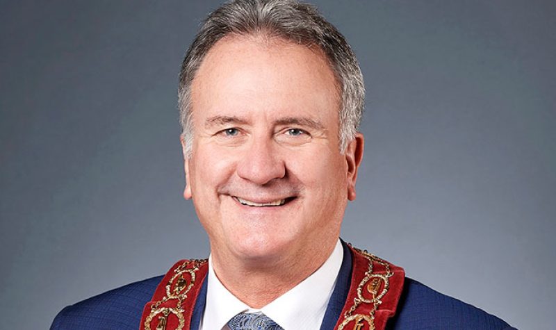 Portrait of Brantford Mayor Kevin Davis. Wearing a red ribbon around suit collar. In front of dark grey backdrop.