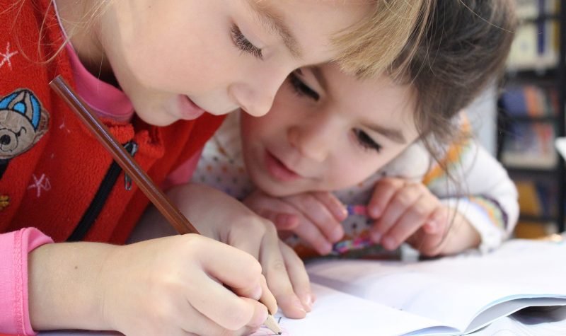 Two children look closely at a book and one of the children is seen writing in the booklet with a colored pencil.