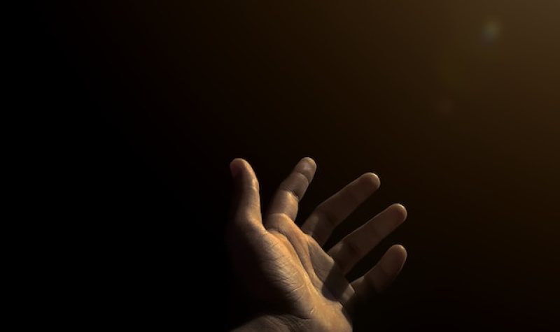 An open  hand reaches out from the bottom of the frame towards the dark. on the right side of the image, a buffered light illuminates the hand