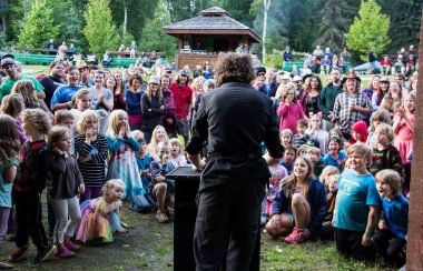 A man's back is to the camera. He is at a podium performing magic for a captive audience of children and adults.