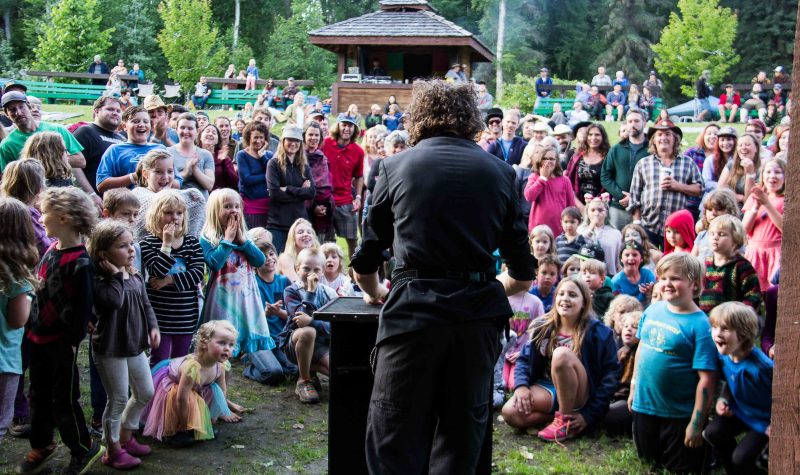 A man's back is to the camera. He is at a podium performing magic for a captive audience of children and adults.