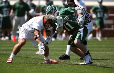 Two men in lacrosse equipment competing for a loose ball. One player in white, other player in green.