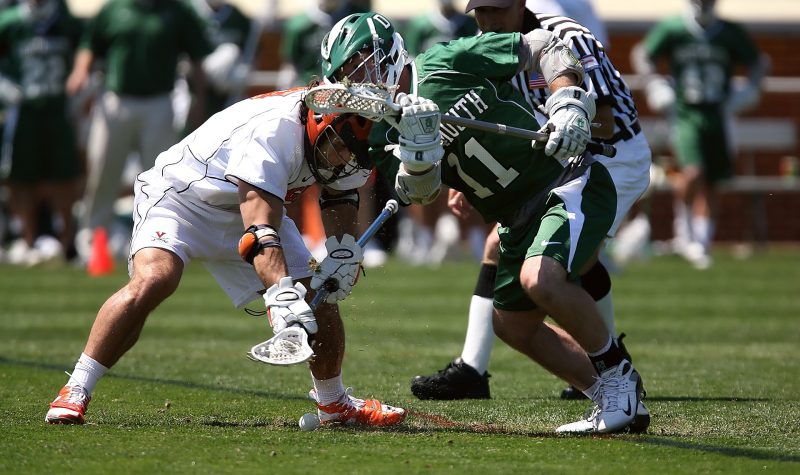 Two men in lacrosse equipment competing for a loose ball. One player in white, other player in green.