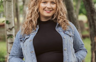 A woman with long hair smiles, standing with hands in jacket pockets.