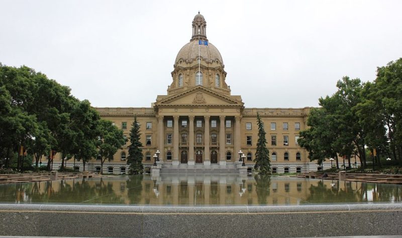 The Alberta Legislature from the front, a tree sits in the photo too, as well as a big pool in front of the building. The Alberta flag waves on top of the building. Weather is partly cloudy.