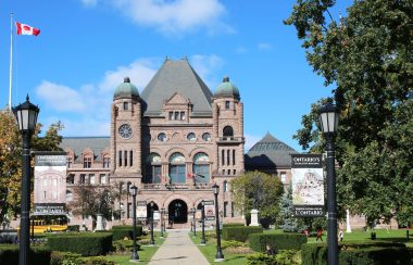 A building with a Canadian flag on the left and trees around during a bright, sunny day.