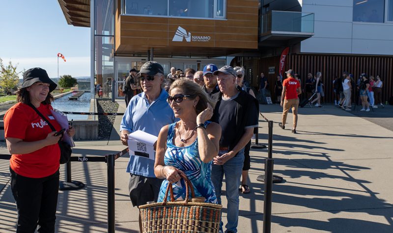 People in a line outdoors on a sunny morning get ready to board the Hullo ferry in front of the Nanaimo Port Authority building.