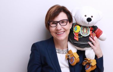Cindy Blackstock stands against a white wall with a teddy bear in Indigenous regalia sitting on her shoulder