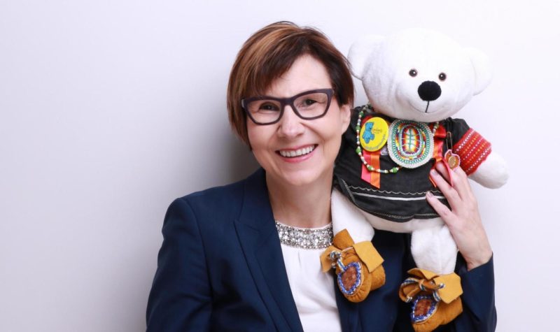 Cindy Blackstock stands against a white wall with a teddy bear in Indigenous regalia sitting on her shoulder