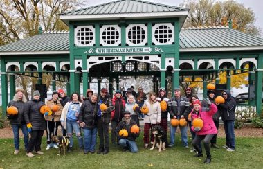 Group gathered in front of green gazebo on grass. Multiple people holding pumpkins.