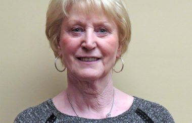 A headshot of Lois Smith, standing in front of a beige background and wearing a grey sweater.