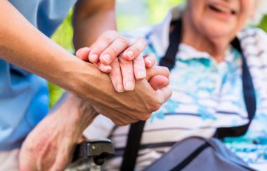 An elderly woman in a wheelchair holds the hand of a healthcare worker in a sunny outdoor environment.