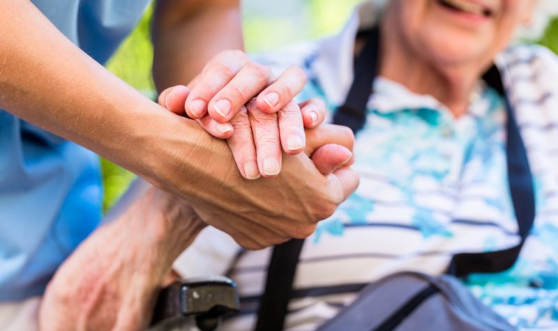 An elderly woman in a wheelchair holds the hand of a healthcare worker in a sunny outdoor environment.