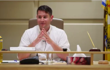 A man in a white shirt sitting at a council desk with hands together in front of him.
