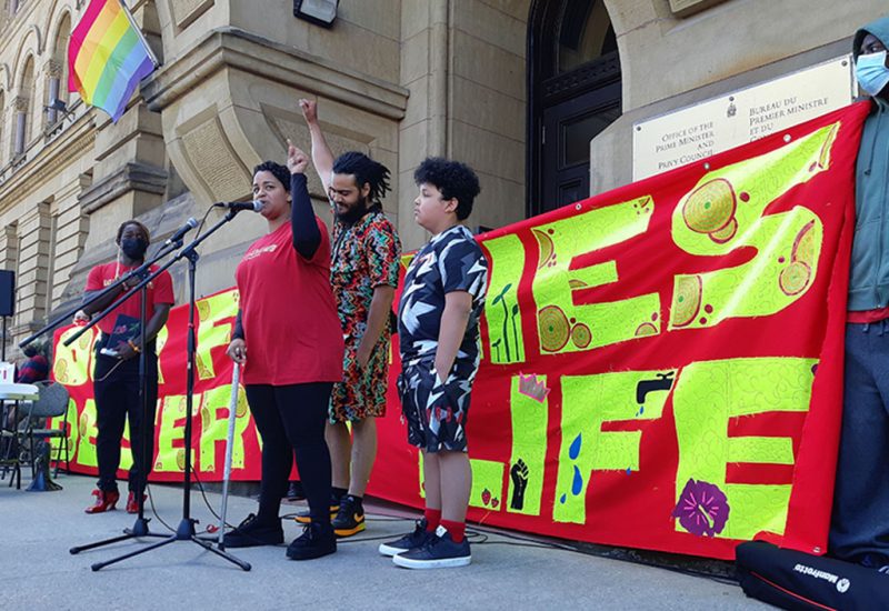 A woman stands at a microphone wo. at the door to the Prime Minister's office. Behind her, a man raises a single fist and there is a red banner that says, 