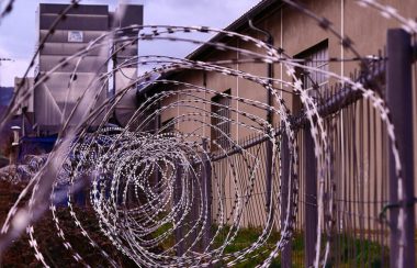 The barbed wire at the top of a fence is pictured. In the background, a penitentiary is seen.