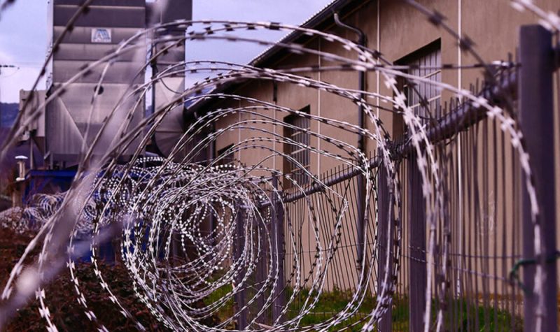 The barbed wire at the top of a fence is pictured. In the background, a penitentiary is seen.