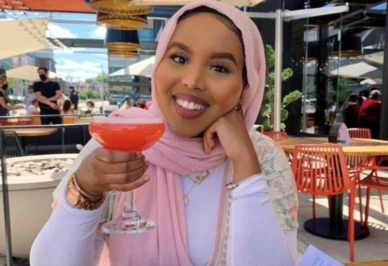 Manal Abdurahman sitting on a patio holding a drink wearing a white long sleeve shirt.