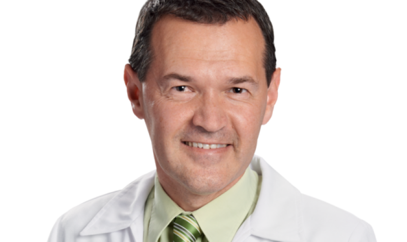 A headshot of pharmacist Marc Aufranc wearing a green dress shirt and tie under a white collared jacket.