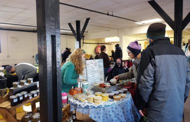 A young woman leans over a table of goods at a farmers market, while a women in a hat looks at an item.