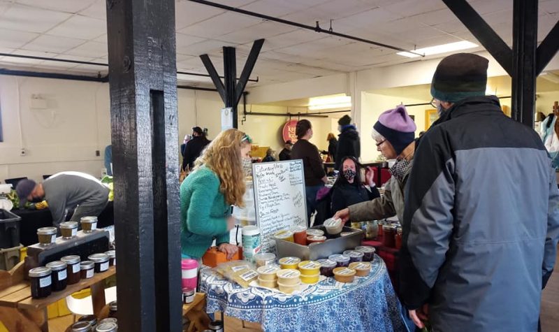 A young woman leans over a table of goods at a farmers market, while a women in a hat looks at an item.