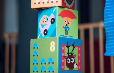 A child's hand steadies the top block of four blocks stacked on top of one another. The blocks have cartoon caricatures of a donkey, cats driving a car, and a moose