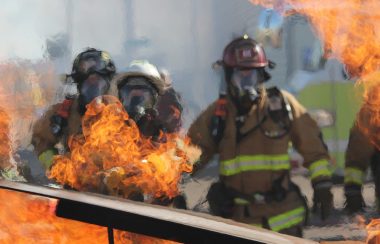 Orange flames on burning debris are shown in the foreground, with firefighters in their protective gear are in the background, the image distorted by heat.