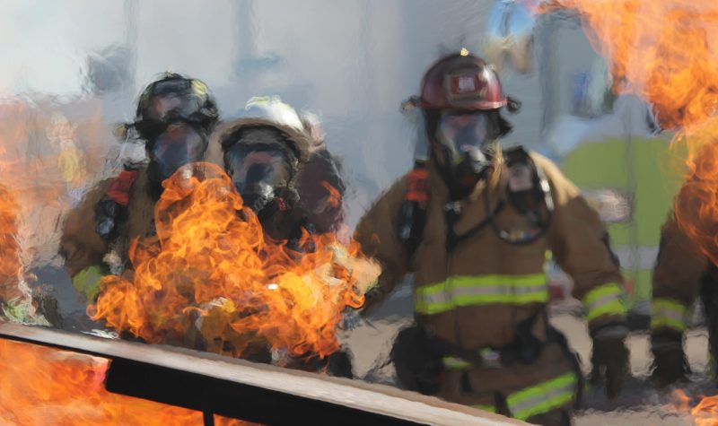 Orange flames on burning debris are shown in the foreground, with firefighters in their protective gear are in the background, the image distorted by heat.
