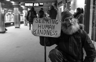 A homeless man wearing a beanie hat and with a bushy grey beard squats on a subway platform holding a sign that reads 'Seeking Human Kindness'. The picture is black and white and there are people blurred in the background