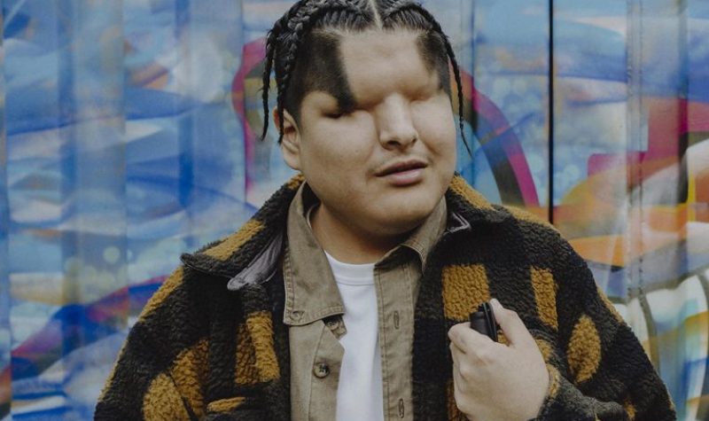 MattAm artist wears a black and yellow plaid jacket. He stands in front of a colorful mural on a corrugated steel surface. He wears a white shirt, holds a cane in his left hand, while the fingers of his left hand also touch the cane. His hair is woven into braids.