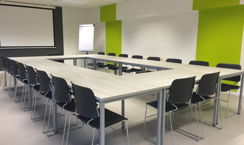 Salle de réunion lumineuse avec plusieurs tables formant un rectangle et nombreuses chaises sombres tout autour.